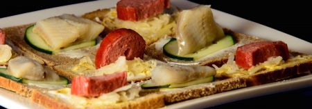 A plate of open-face Danish-style sandwiches, "Tak Snacks" as seen from the side so you can see the sausage and herring standing up like standing stones on the Tak board.